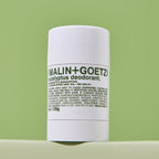 A white cylindrical bottle of Malin+Goetz Eucalyptus Deodorant standing upright against a light green background.