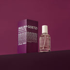 A bottle of (Malin + Goetz) Moroccan Fig perfume next to its purple packaging box, set against a matching purple background.